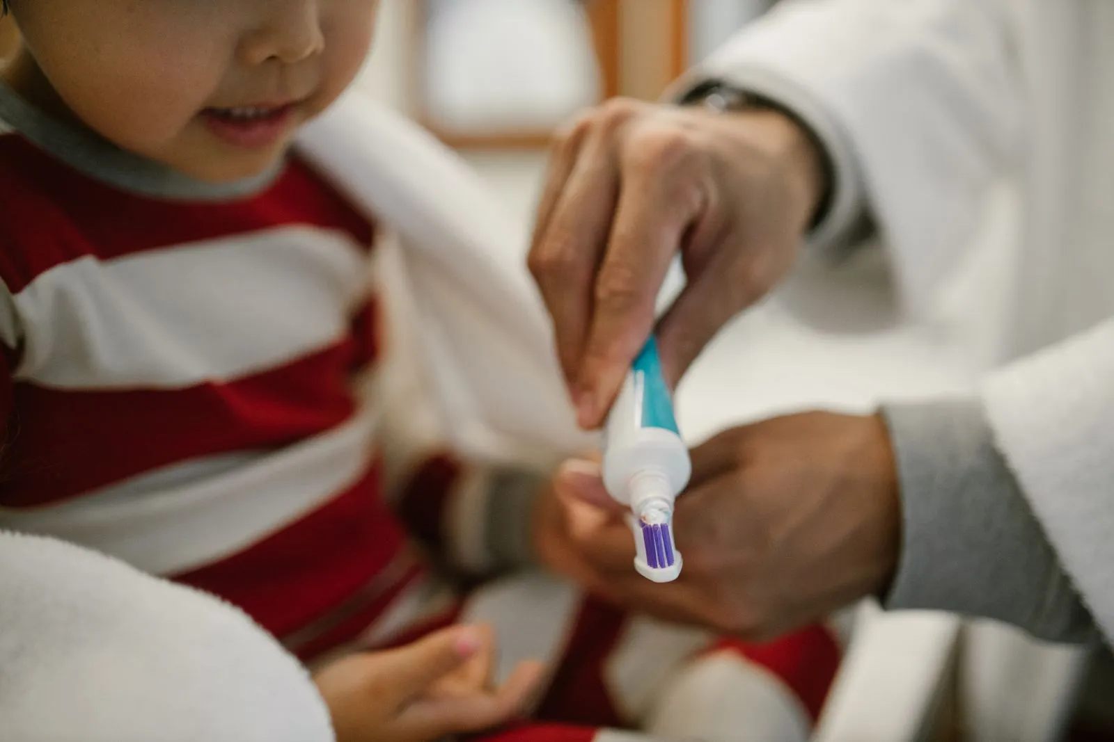A close-up of a father helping his young daughter apply toothpaste on a toothbrush.
